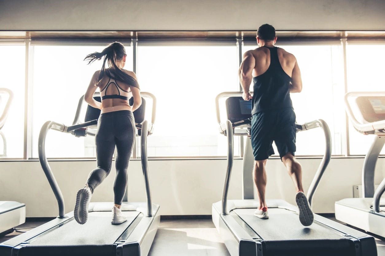 Two people are running on a treadmill in front of windows.